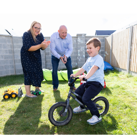 mother and father look at young son on his bike in back garden, with grey brick wall in background, grass, wooden fencing and toys in the background