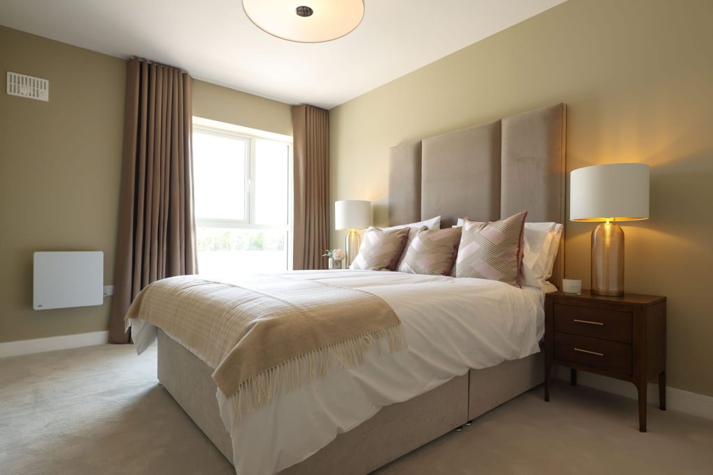 Show home double bedroom at Bennetstown with an upholstered headboard, bedside lamps and a curtained window.