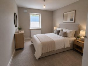 Show home double bedroom at Hearthfield with neutral bedding, a bedside lamp, a chest of drawers with a round mirror, and a door opening onto the landing.