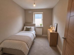 Show home single bedroom at Hearthfield with a single bed, a bedside lamp, a chest of drawers and a window above the radiator.